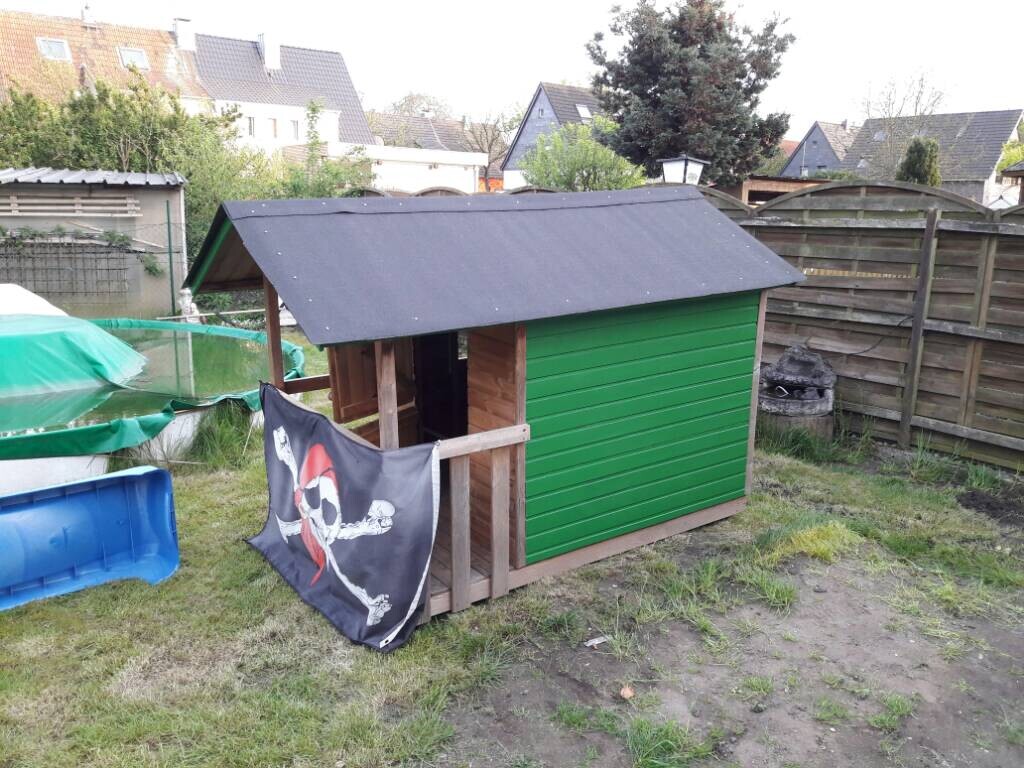 Kleines grünes Gartenhaus mit schwarzem Dach im Garten, Piratenflagge davor