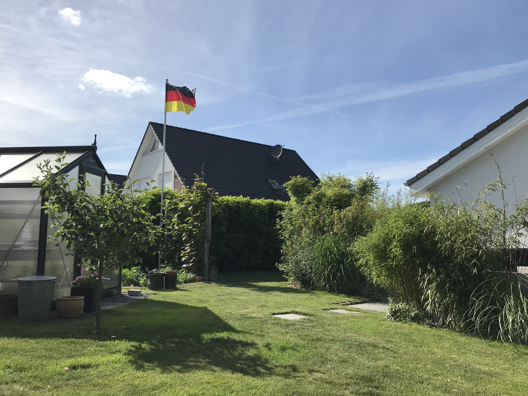 Garten mit grünem Rasen, Hecke, Flagge Deutschlands auf Mast, Gewächshaus links.