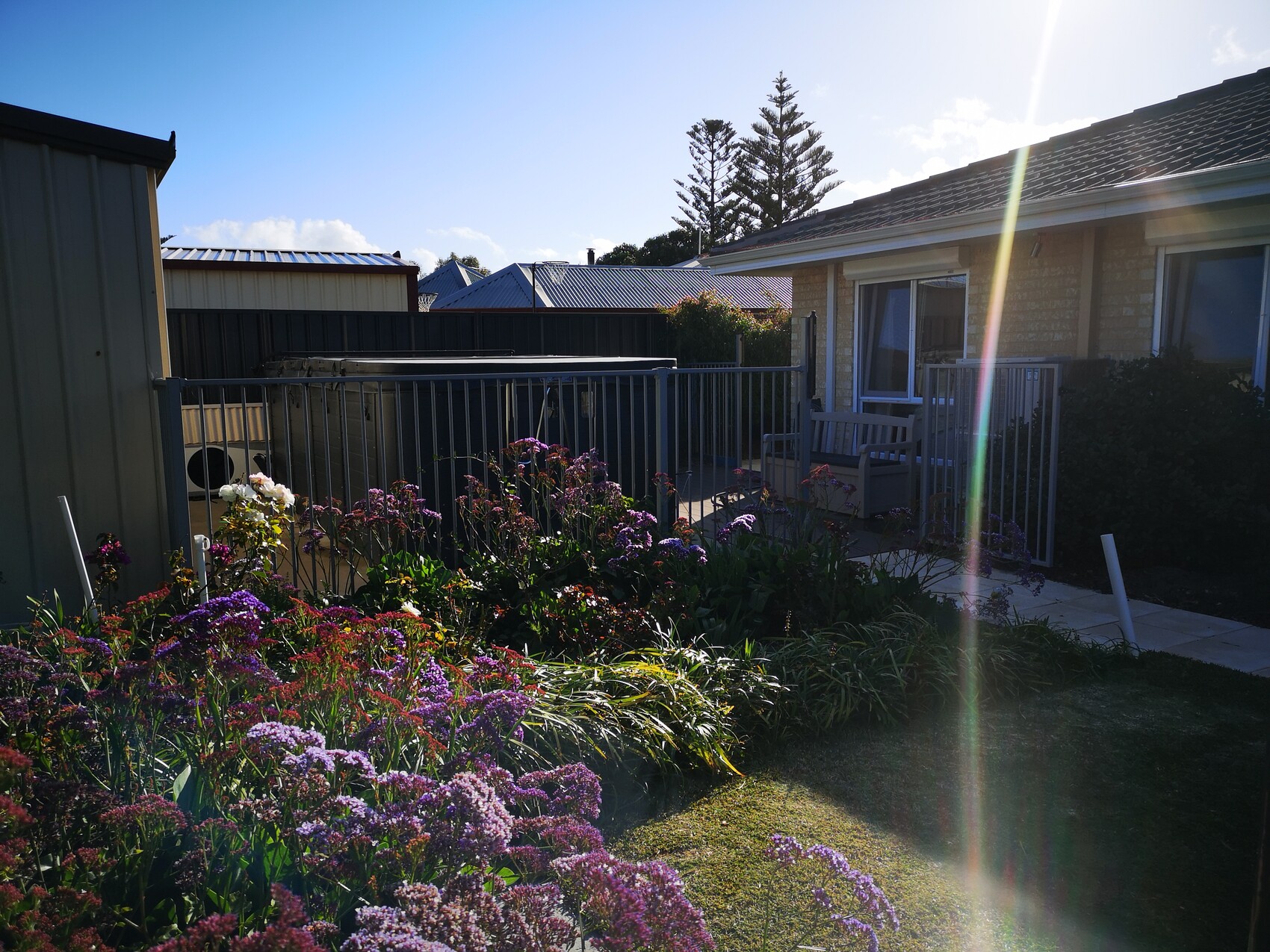 Blumenbeet im Vordergrund, Haus mit Veranda hinter Metallzaun; Gartenmöbel und Sonnenstrahlen rechts.