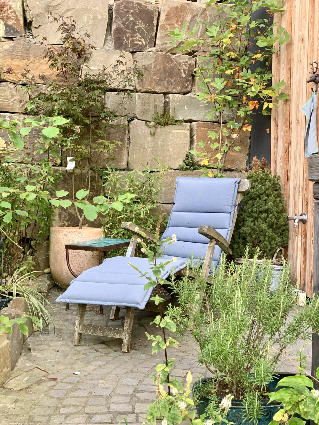 Garten-Lounge mit blau gepolsterter Sonnenliege vor Steinmauer, von Pflanzen umgeben.