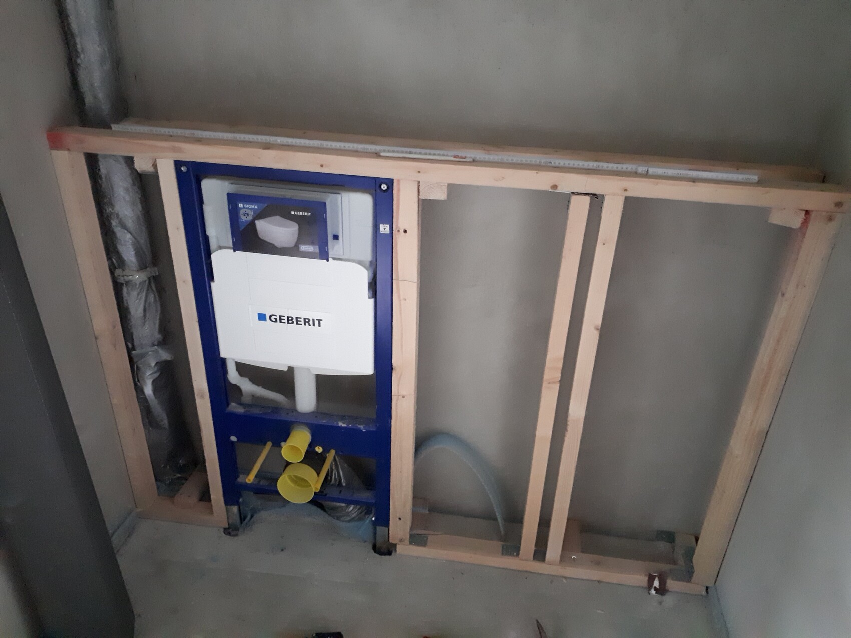 Geberit Spülkasten-Wandinstallation hinter Holzständerwerk, sichtbare Rohre und Bauteile.