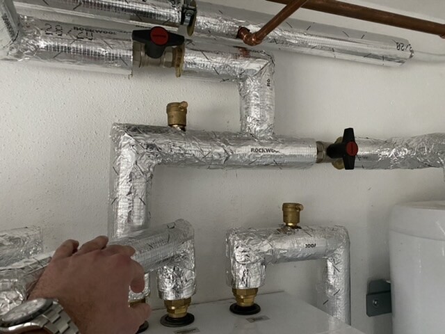 Isolierte Metallrohrleitungen mit Ventilen an einer Heizungsanlage; eine Hand bedient einen Hahn.