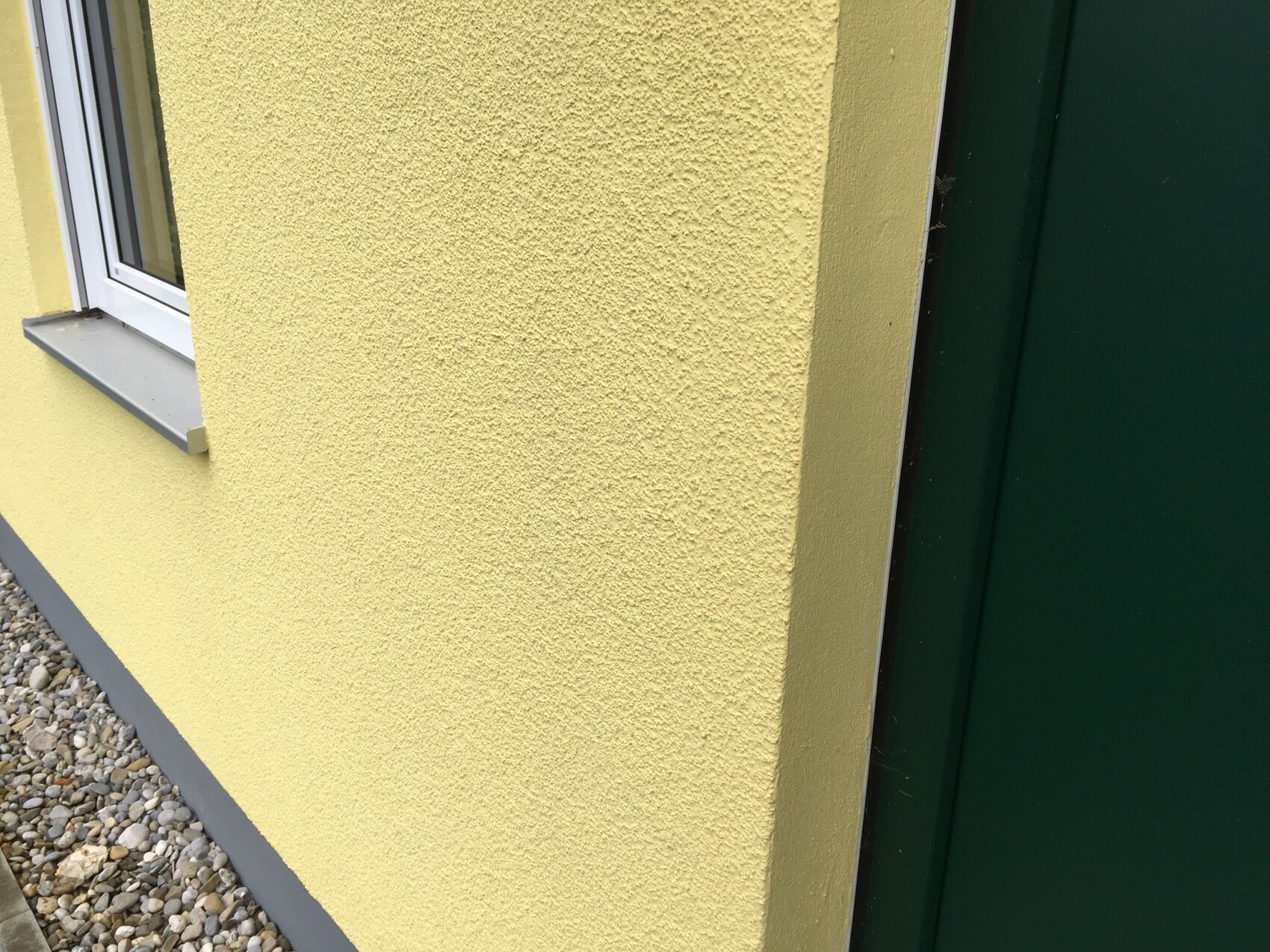 Außenwand: gelbe grobe Putzstruktur, Fenster links, grüne Tür rechts, Kiesboden.
