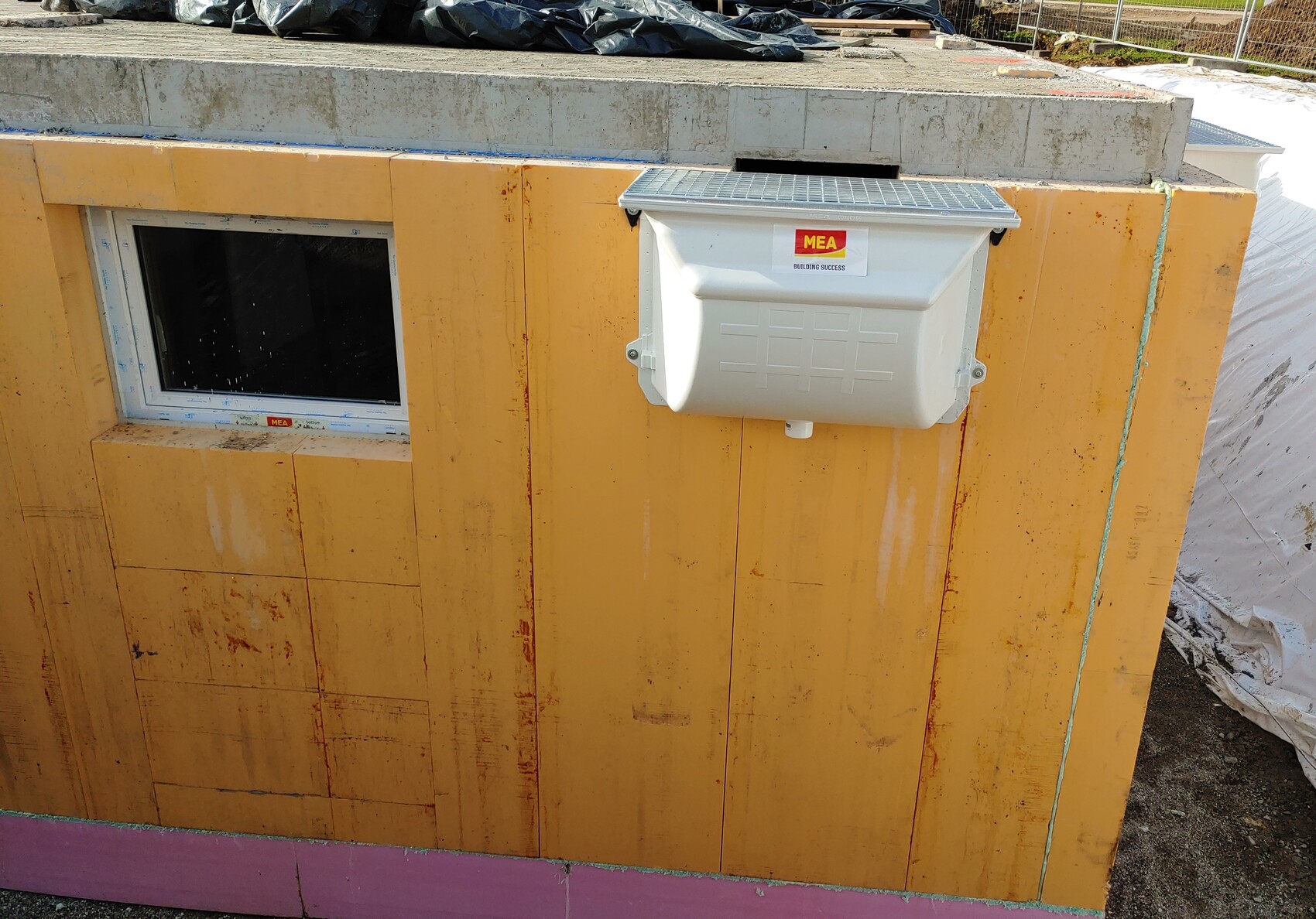 Baustellenwand in gelber Verkleidung mit kleinem Fenster links und weißer MEA-Verteilerbox rechts.