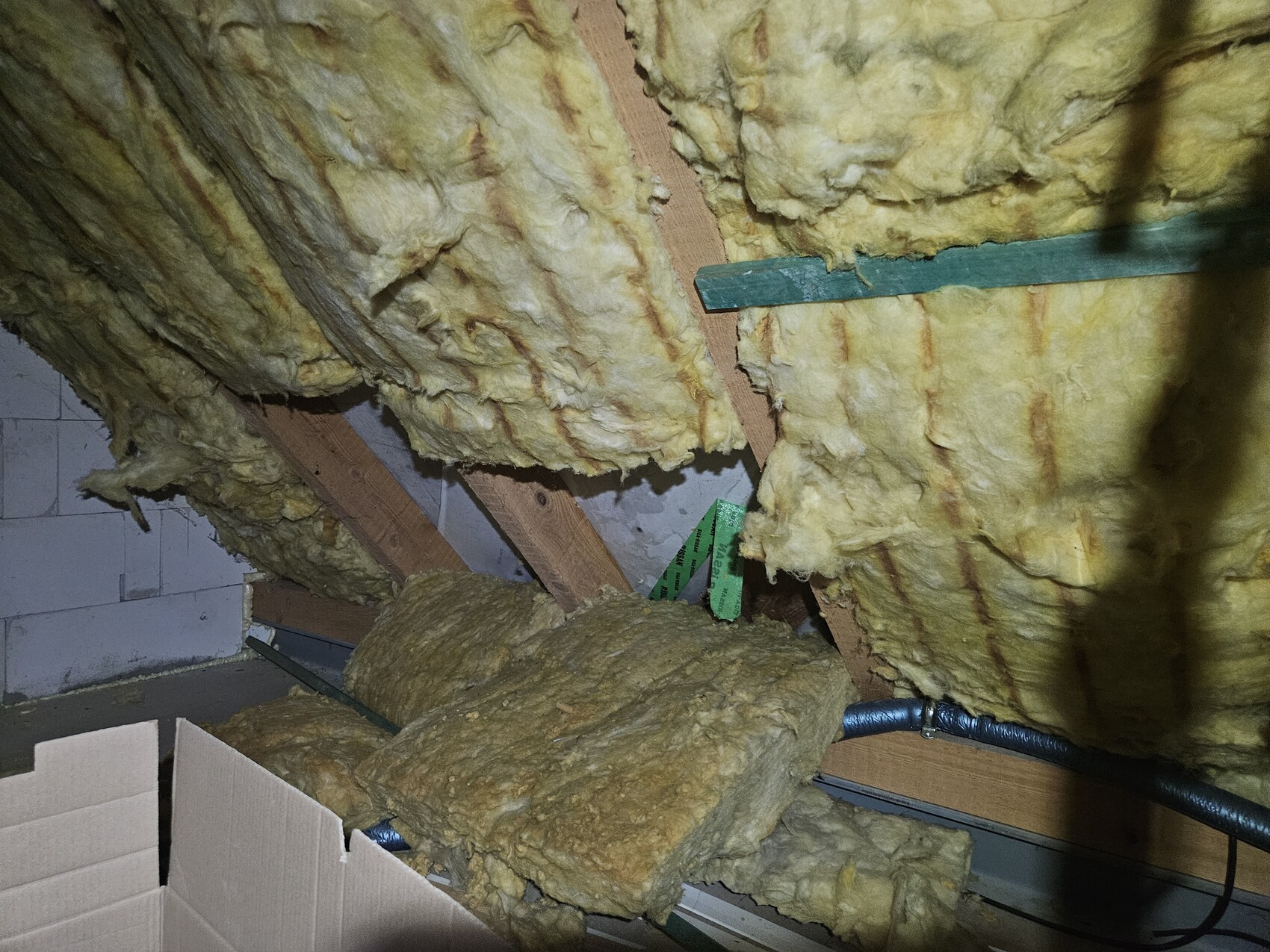 Dachboden: gelbe Dämmwolle zwischen Holzbalken, Kartonbox links, sichtbare Rohrleitungen unten.