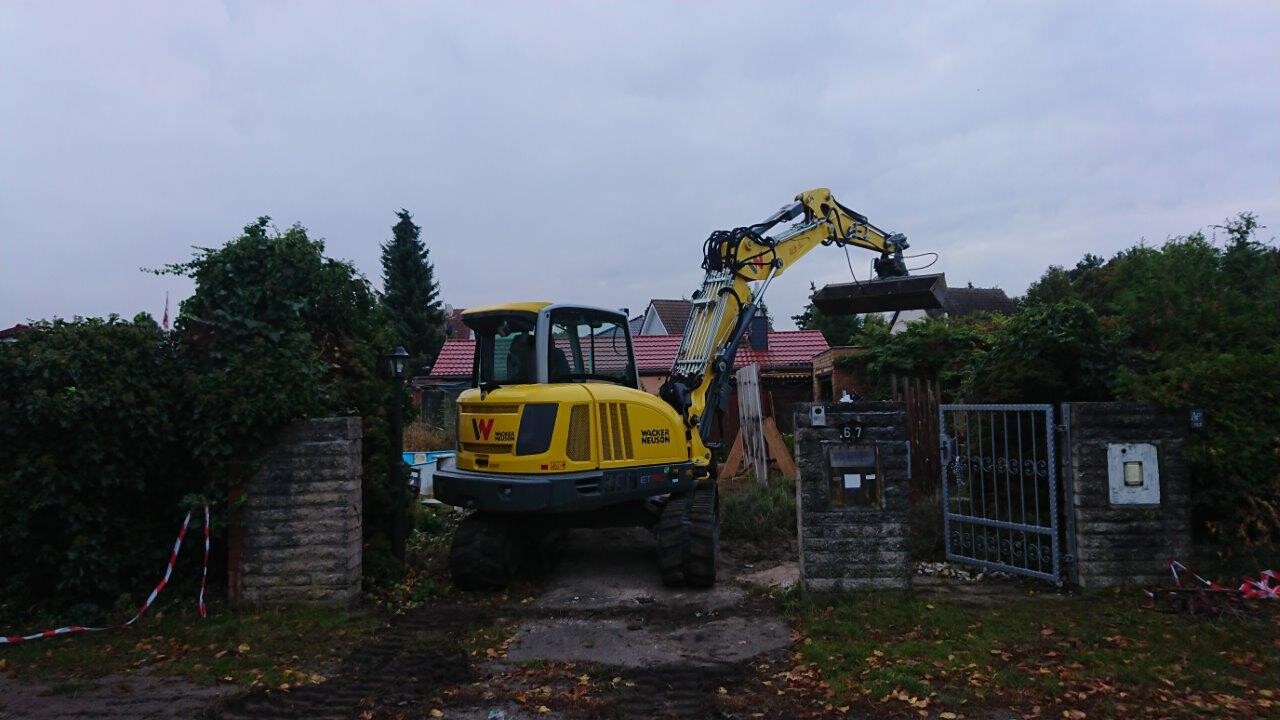 Gelber Bagger mit ausgestrecktem Arm vor Garteneinfahrt; Bauarbeiten im Vorgarten.