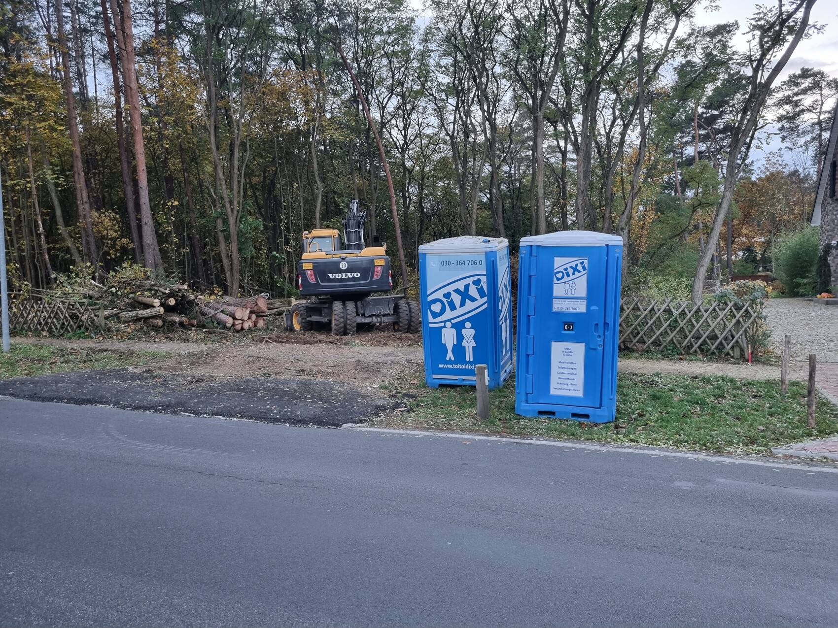 Baustelle mit Volvo-Erdbaumaschine, Holzstapel links, zwei blaue DIXI-Toiletten rechts.