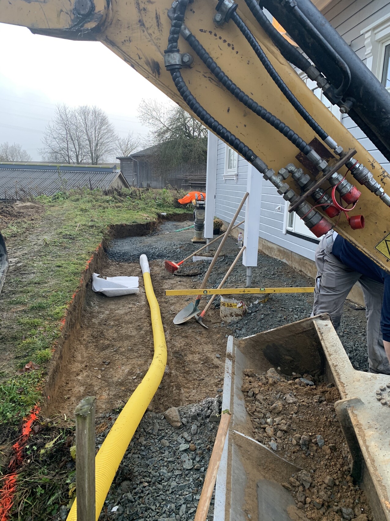 Baustelle neben Haus: Baggerarm, gelber Schlauch führt durch Graben; Schaufeln und Werkzeug.