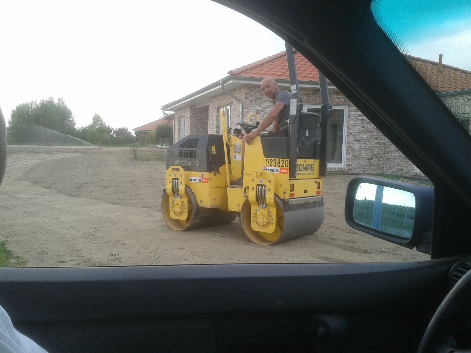 Gelber Bauroller mit Fahrer auf Baustelle vor Einfamilienhaus, Sicht aus Autofenster