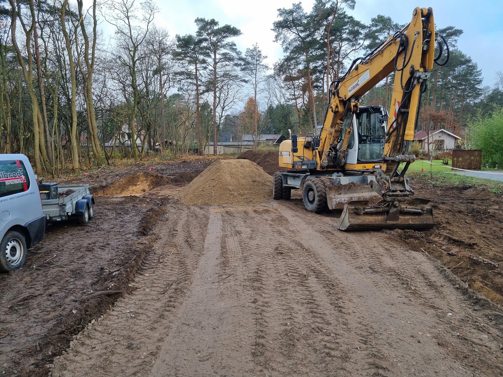 Gelber Bagger auf unbefestigter Erdstraße, Erdhaufen und Wald im Hintergrund.