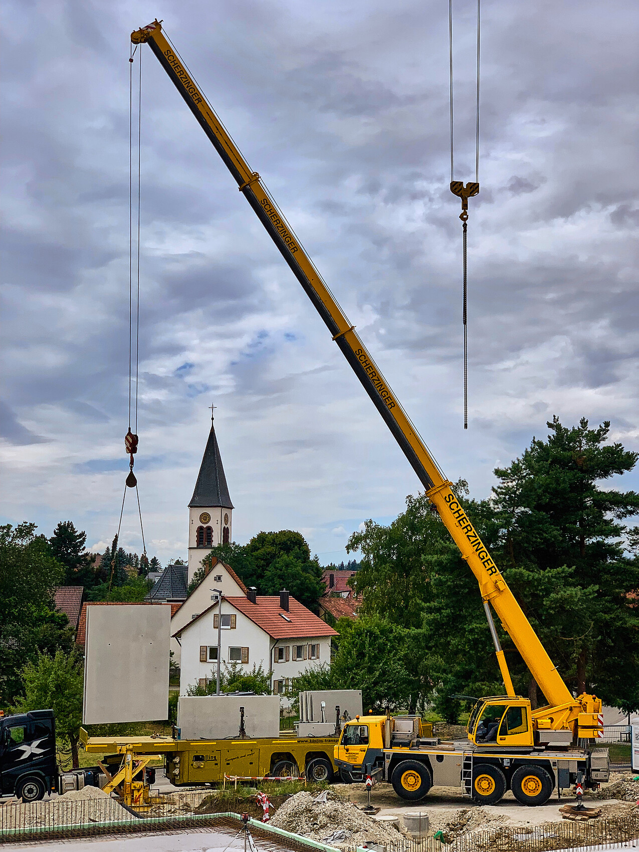 Großer gelber Mobilkran hebt Betonplatte auf Baustelle; Kirchturm und Häuser im Hintergrund.