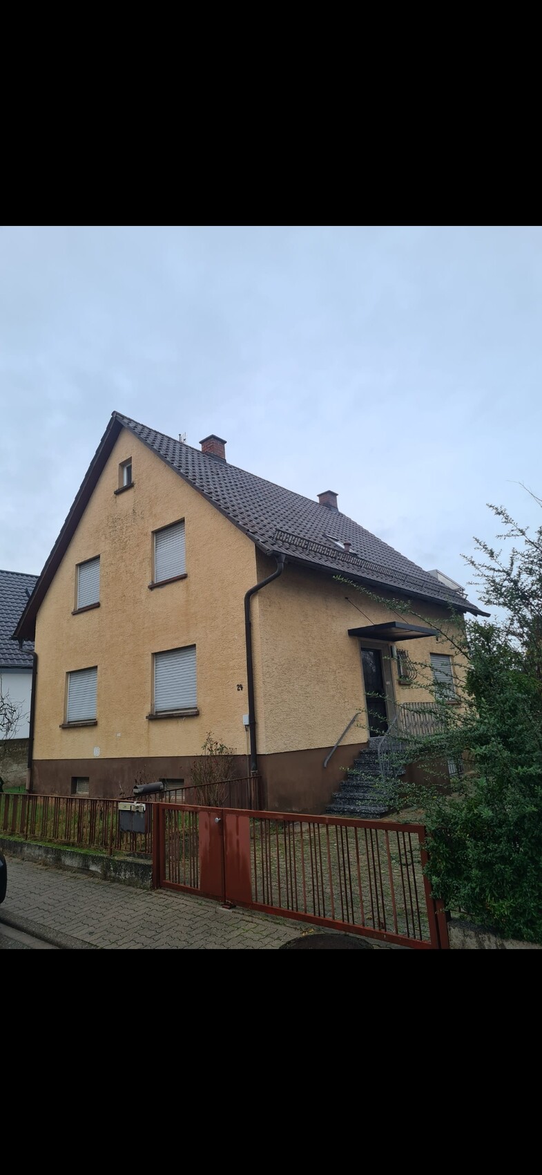 Zweistöckiges Haus mit gelber Fassade, grauem Dach, Treppe zum Eingang und rotem Zaun.