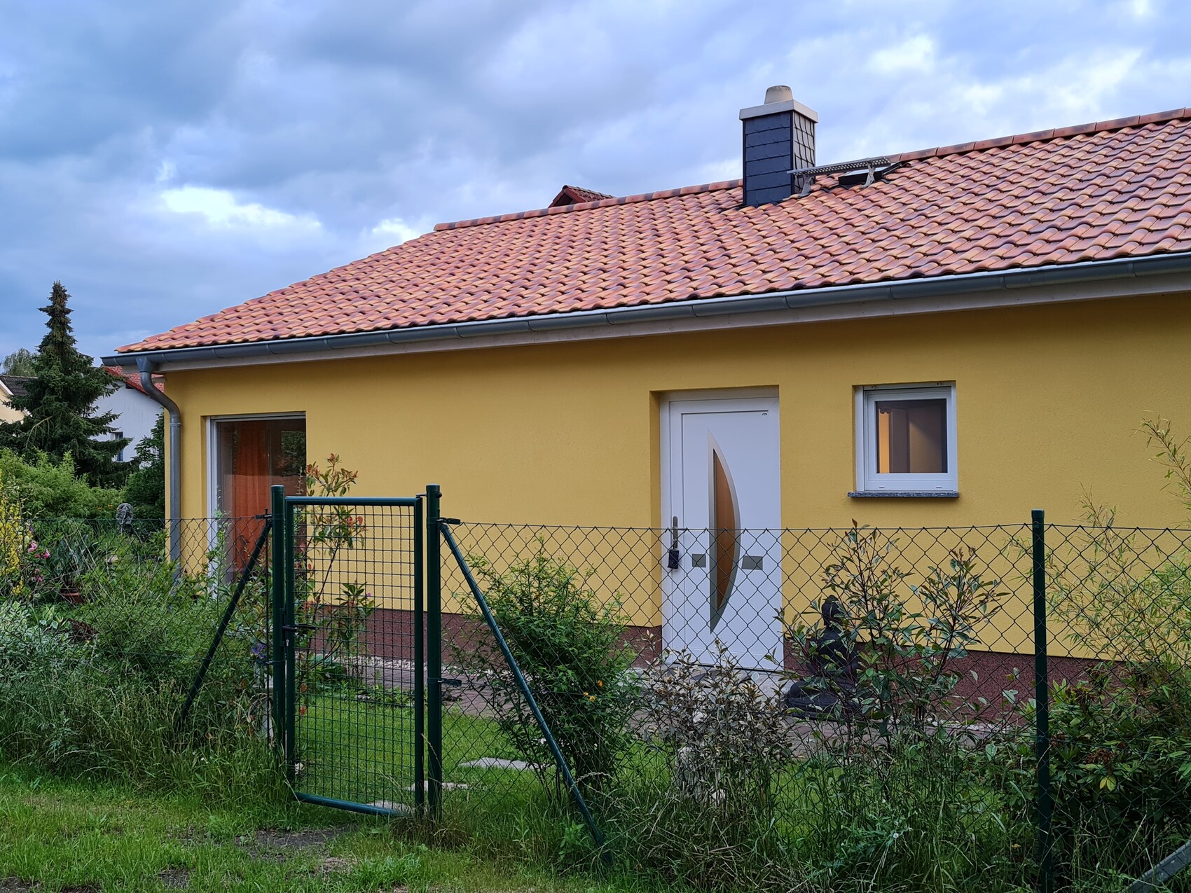 Gelbes Einfamilienhaus mit rotem Ziegeldach, weißer Tür mit Glasfenster und grünem Maschendrahtzaun.