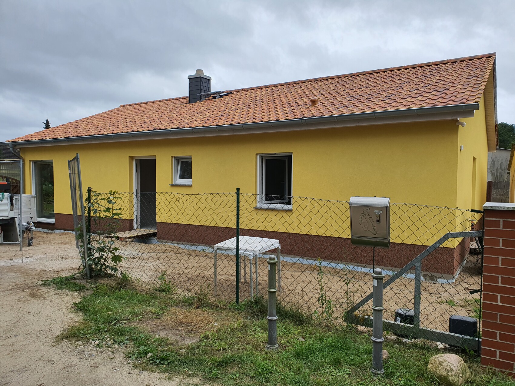 Gelbes Einfamilienhaus mit rotem Sockel und Ziegeldach, Zaun, Briefkasten im Hof.