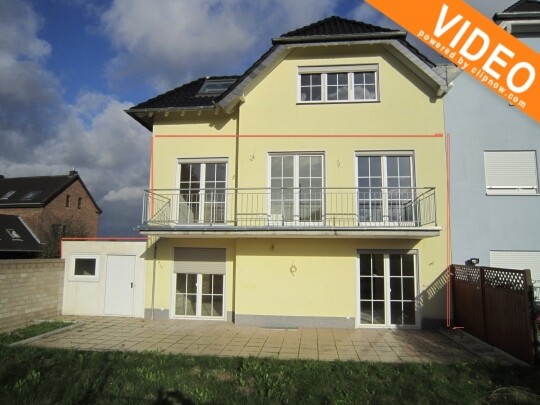 Zweigeschossiges gelbes Einfamilienhaus mit Balkon, Terrasse und Garten vor blauem Himmel.