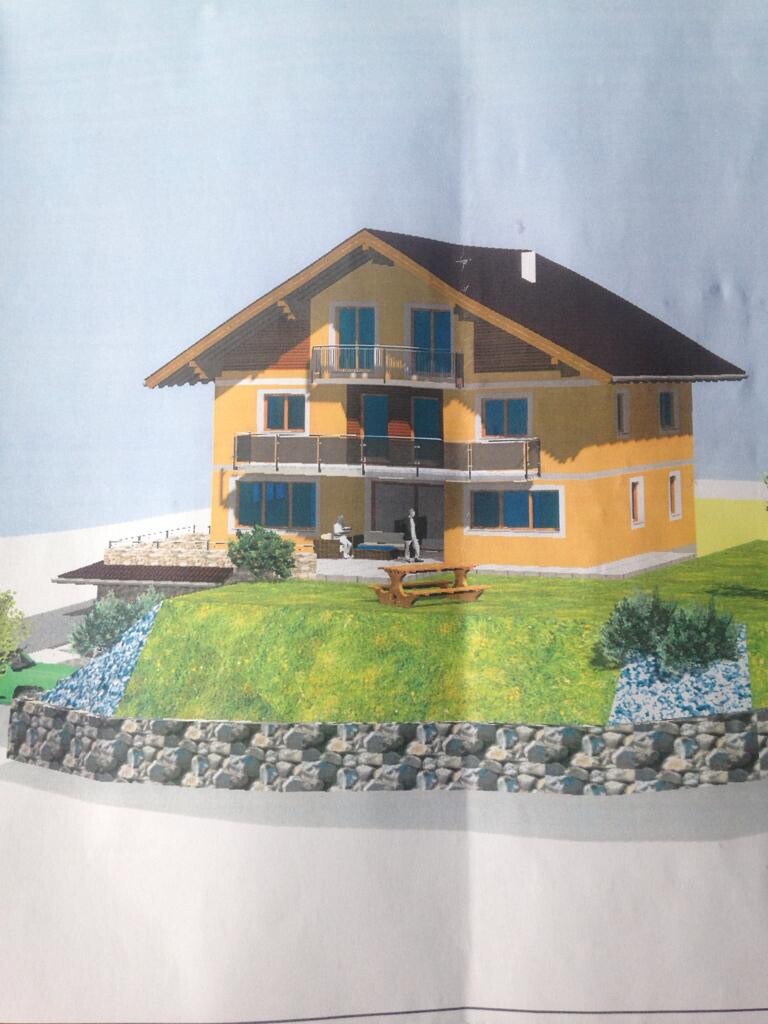 Modell eines gelben, zweistöckigen Hauses mit Balkon und Terrasse auf grünem Hang mit Steinmauer.