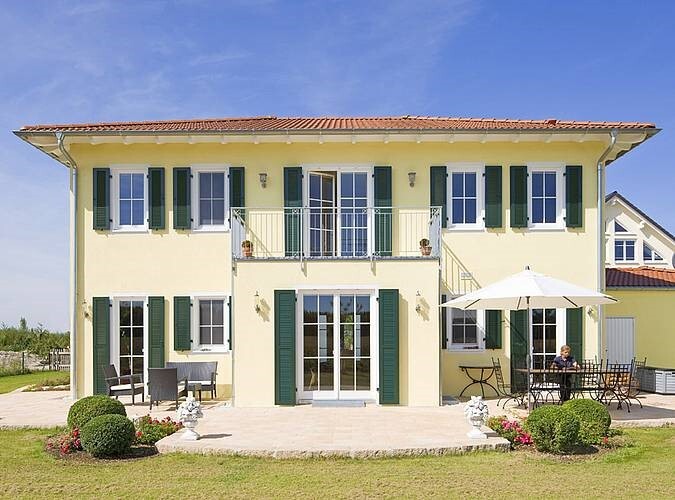 Helles Zweifamilienhaus mit grünen Fensterläden, Balkon, Terrasse und Personen draußen.