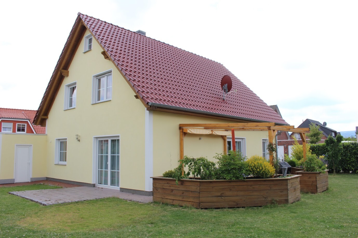 Gelbes Einfamilienhaus mit rotem Ziegeldach, Terrasse und bepflanzten Holzkübeln im Garten.
