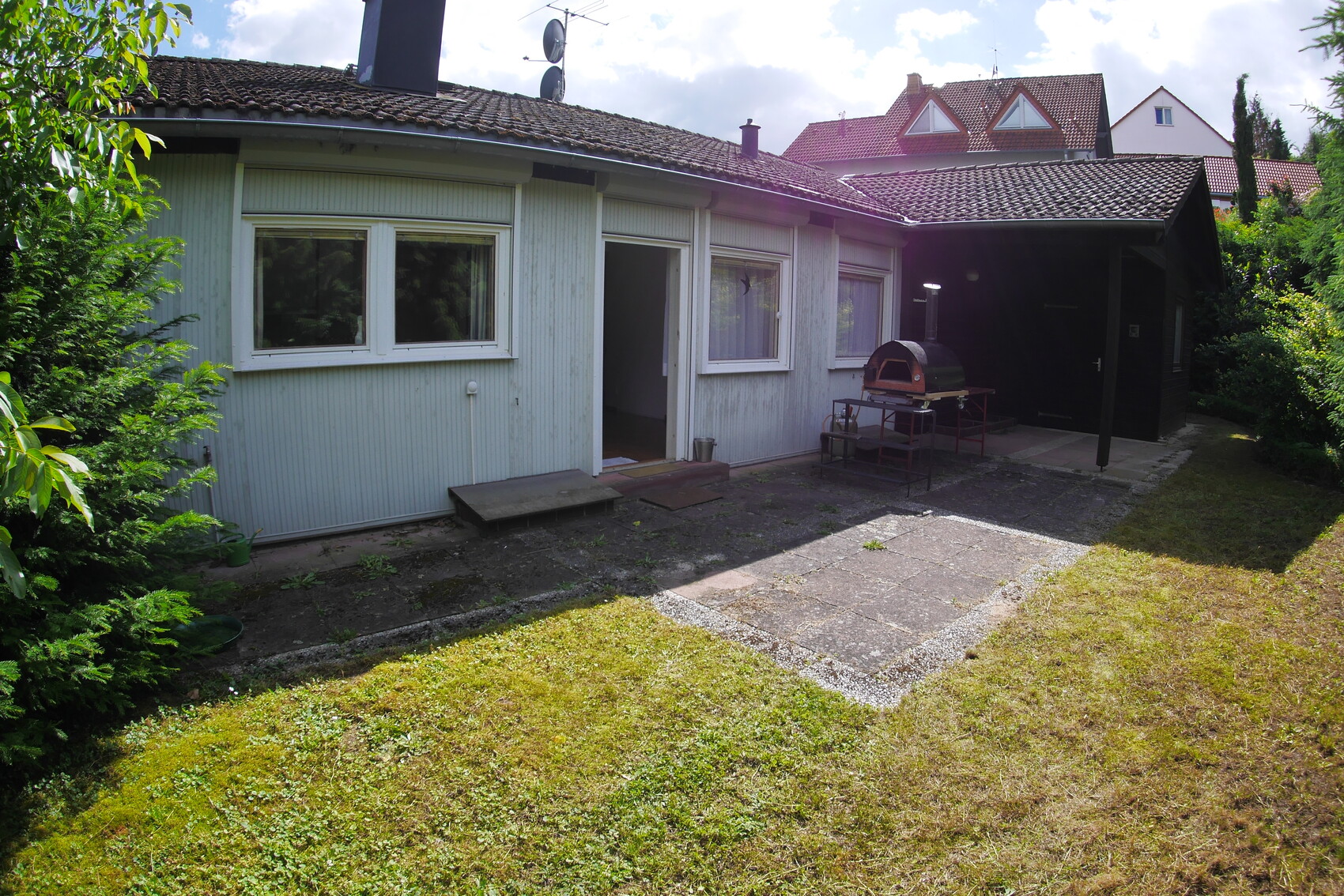 Hinterhof eines Hauses: Terrasse mit Grill, Tür, Fenster, Rasenfläche und Hecken.