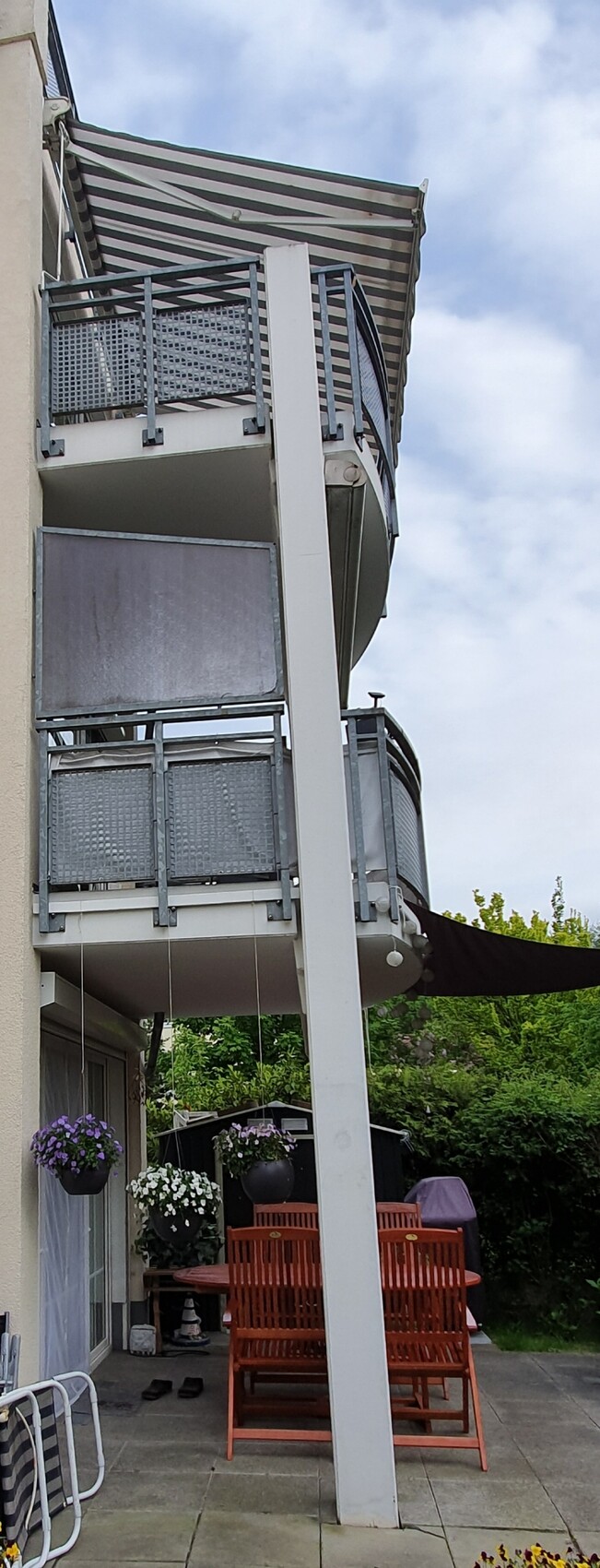 Balkon mit Metallgeländer, gestreifte Markise, Outdoor-Tisch, Stühle und Blumenkübel.