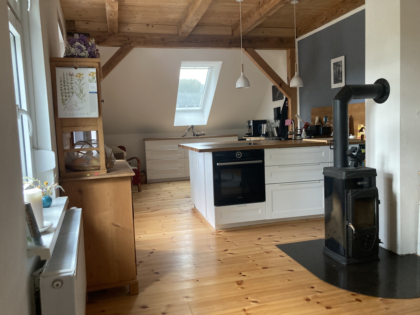 Küche im Dachgeschoss mit Holzbalken, Holzfußboden, Inselküche, Kaminofen und Dachfenster.