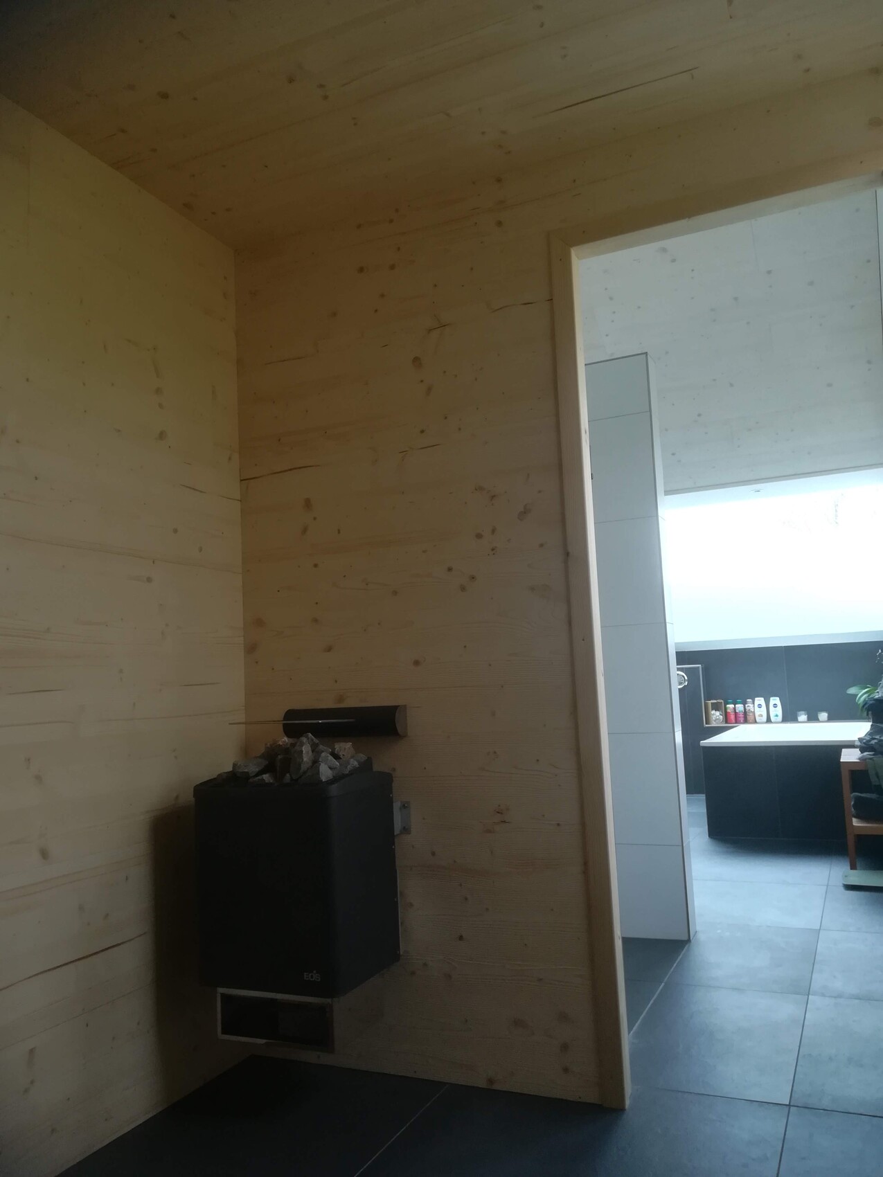 Holzverkleideter Raum mit Saunaofen und Steinauflage; Blick in angrenzende Küchenecke mit Fliesen.