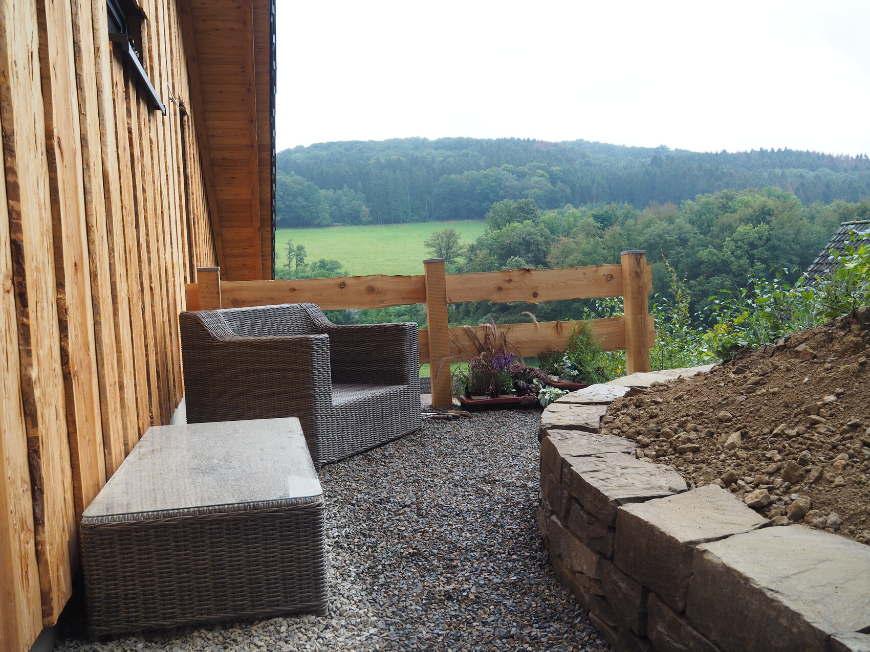 Holzveranda mit Rattan-Sessel, Stein-Tisch, Pflanzkübeln; Blick auf Felder und Wald.