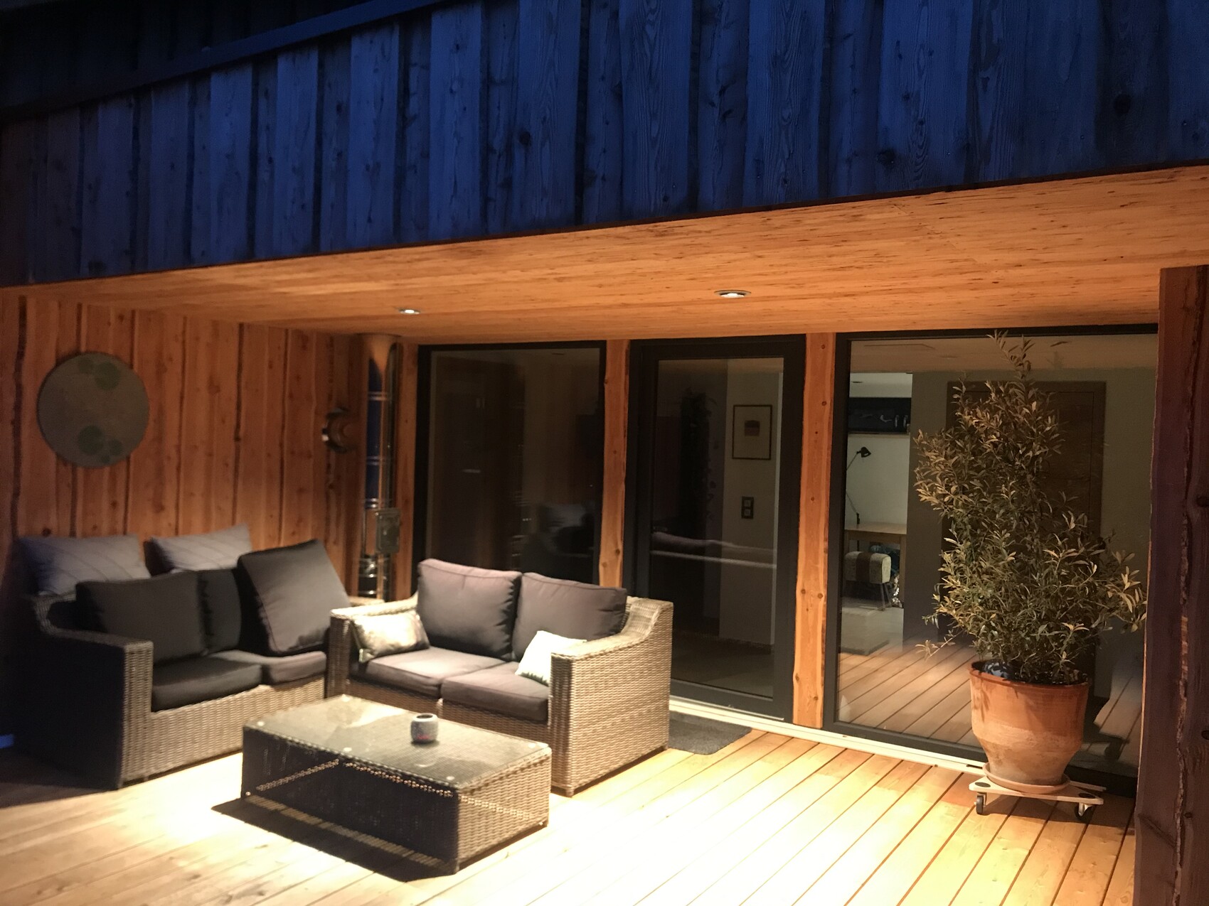 Gemütliche Holzterrasse mit grauer Couch-Gruppe, Glas-Couchtisch und Pflanze vor Glas-Schiebetüren.