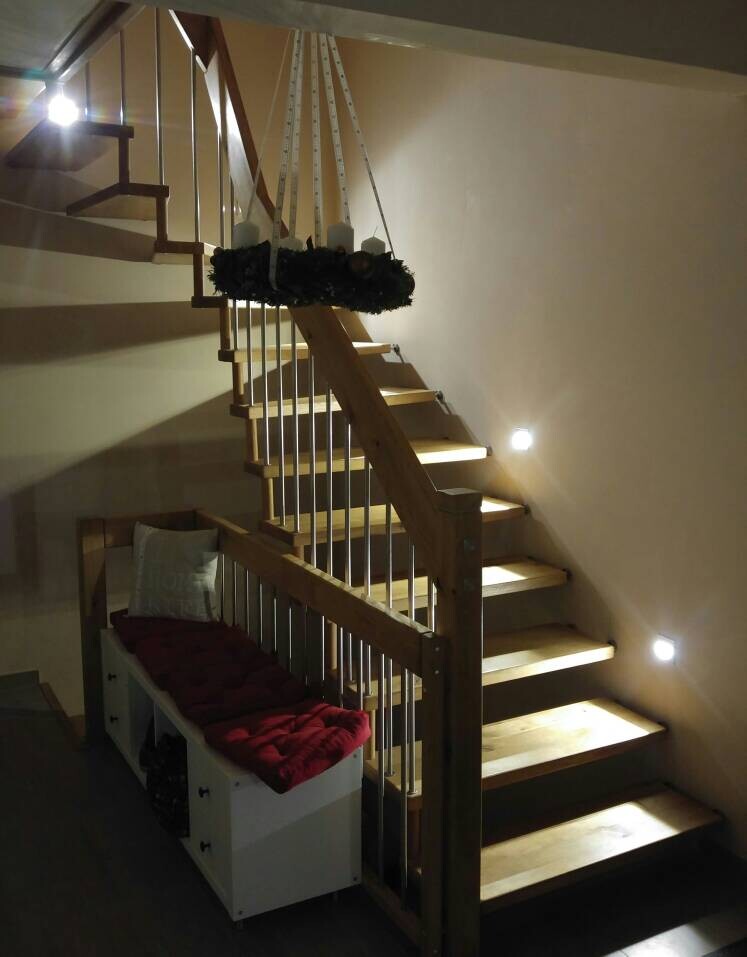 Holztreppe mit Geländer; ein dunkler Kranz hängt über den Stufen, unten rote Kissen auf der Bank.