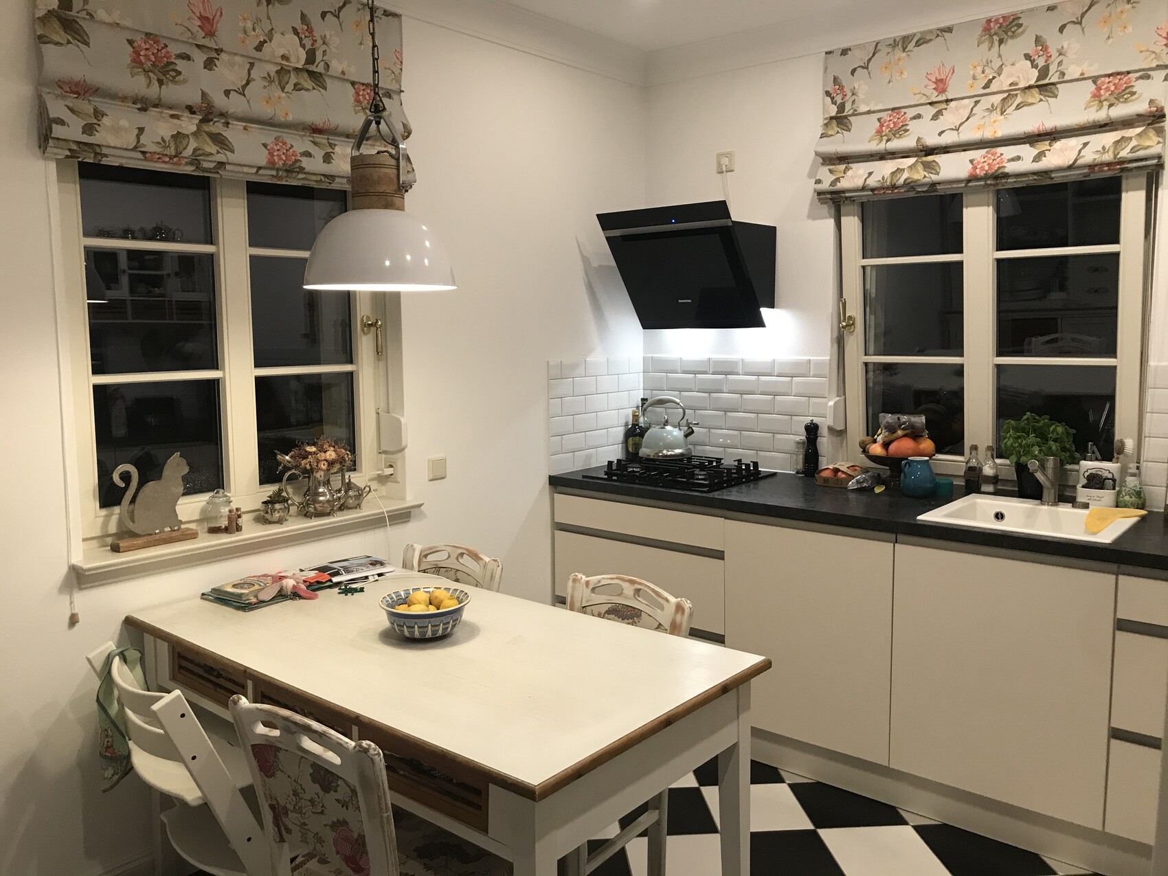 Helle Küche mit weißen Schränken, Fenster mit Blumen-Vorhang, Spüle und Esstisch mit Zitronen.