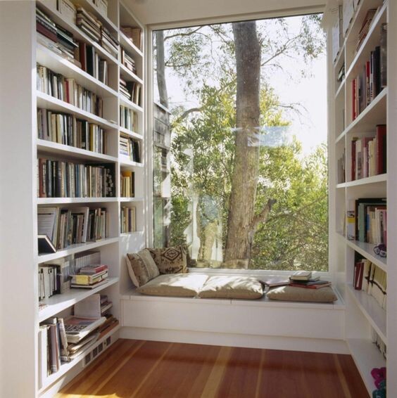 Helle Leseecke zwischen Bücherregalen, großes Fenster mit Blick auf Bäume, Sitzbank.