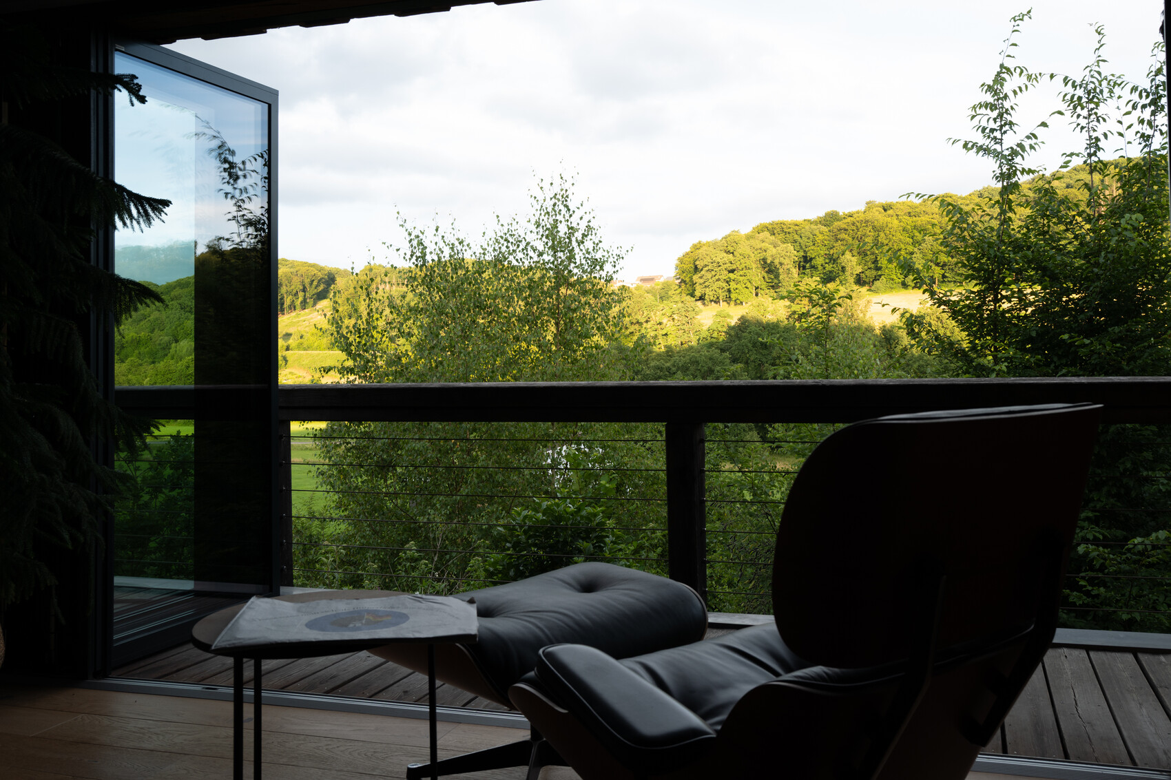 Offene Balkontür, dunkler Lounge-Sessel mit Fußstütze, Tisch mit Magazin, Blick auf grüne Hügel.