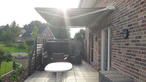 Gepflasterte Terrasse vor Backsteinhaus mit ausgefahrener Markise, runder Tisch und Sitzgruppe.