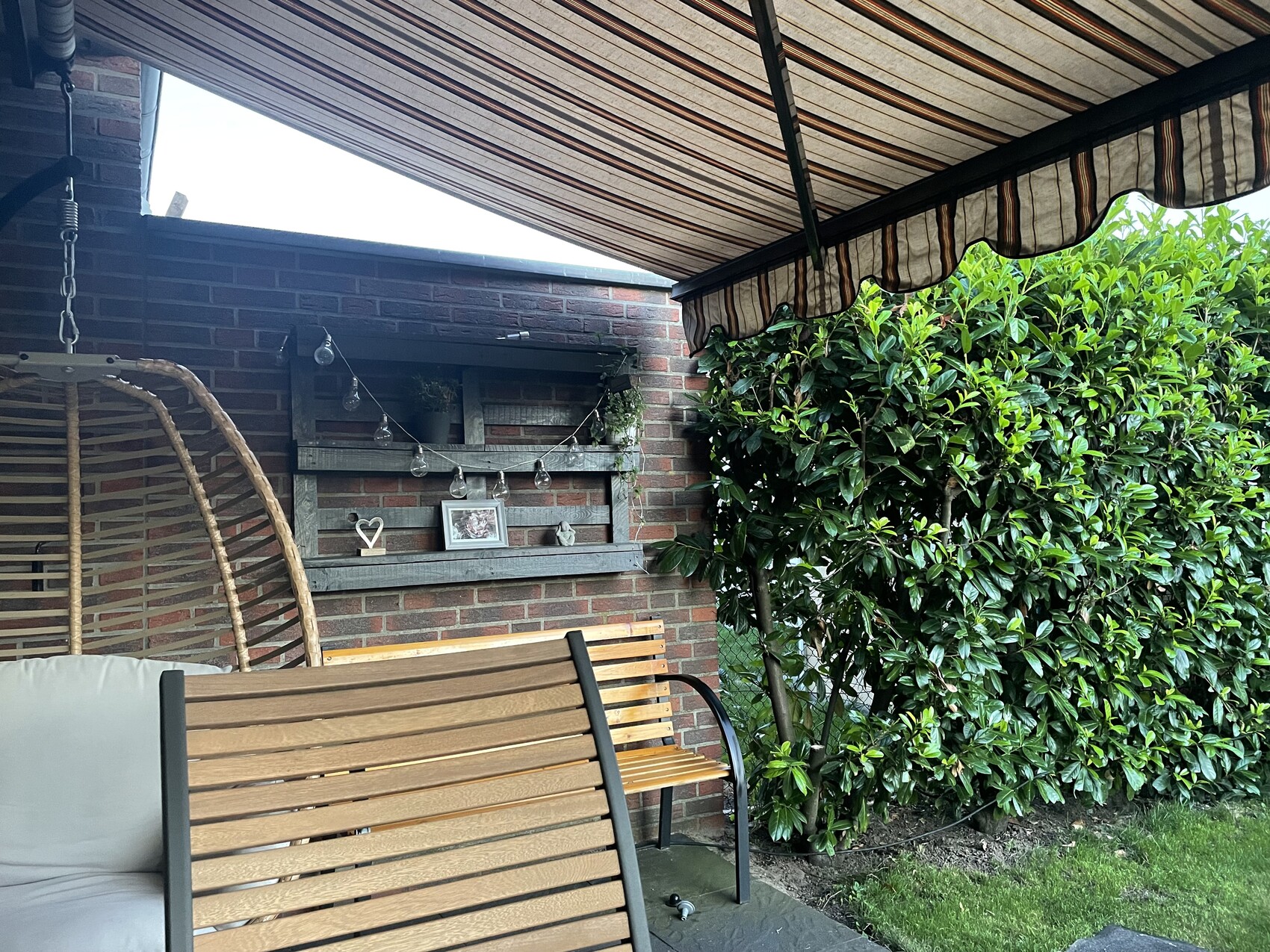 Gemütliche Terrasse mit Holzbänken, Hängesessel, Lichterkette, Deko-Regal an Ziegelwand, Hecke.