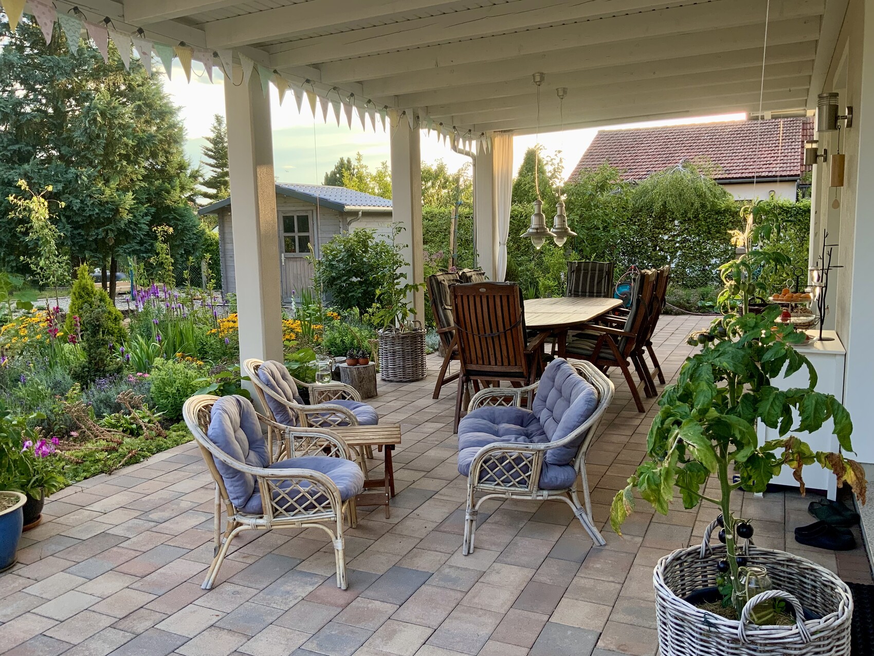 Überdachte Terrasse mit Esstisch, Rattanmöbeln, Blumenbeeten und Gartenpflanzen.