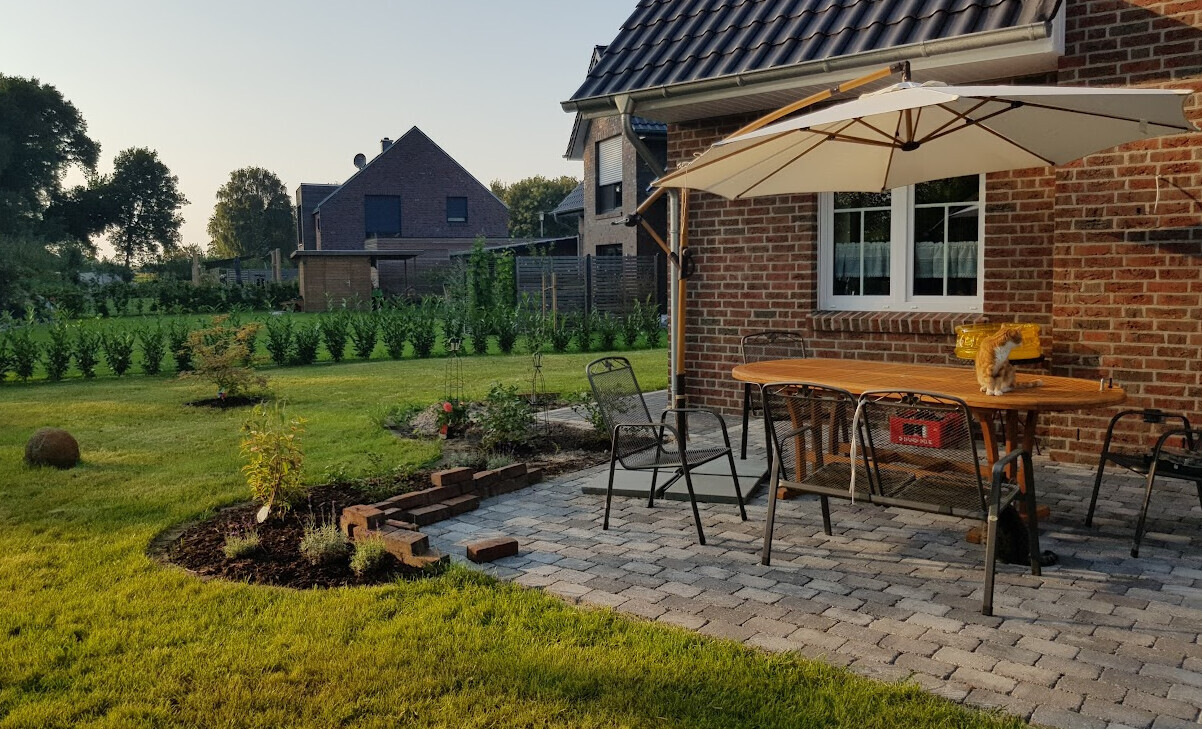 Garten-Terrasse vor Backsteinhaus mit Tisch, Stühlen und Sonnenschirm; Katze auf dem Tisch.
