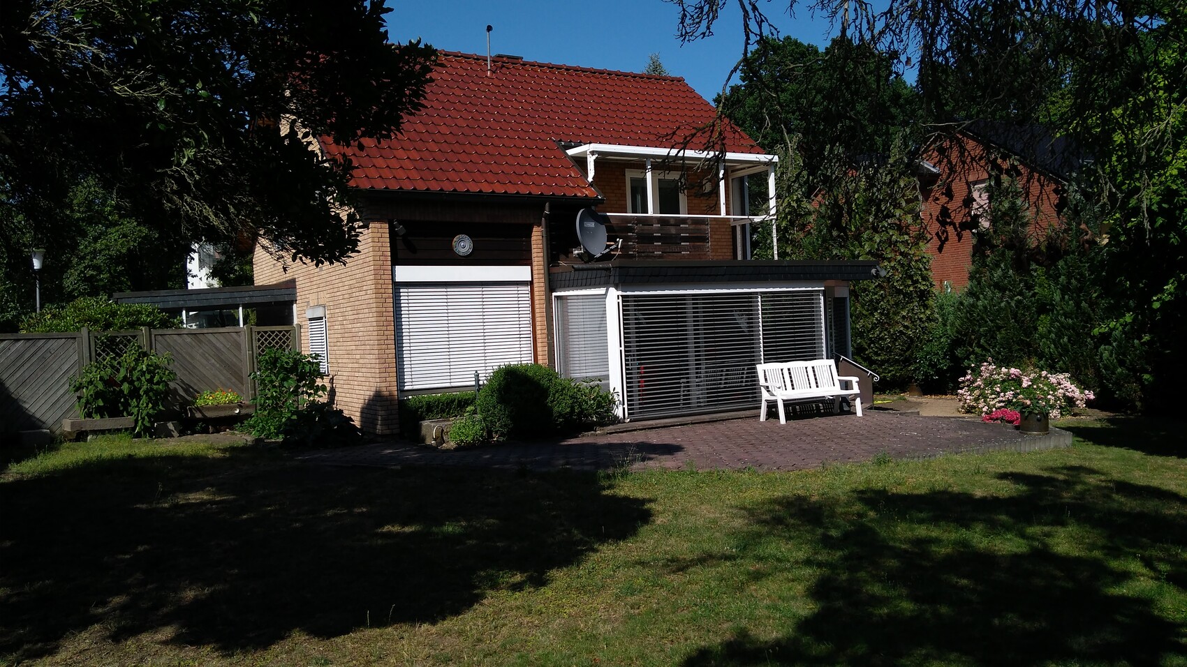 Zweistöckiges Haus mit rotem Ziegeldach, Glas-Überdachung, Balkon und weißer Gartenbank im Garten.