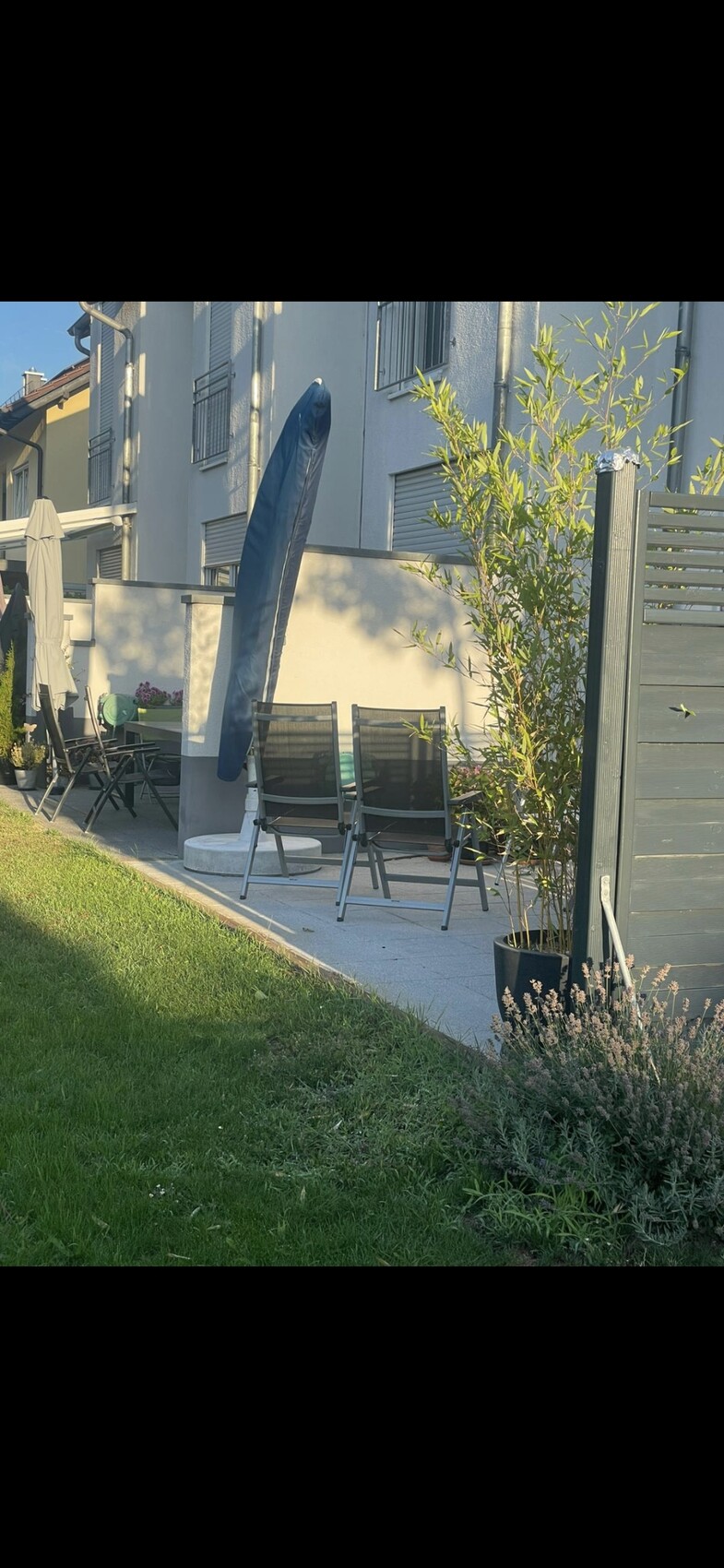 Garten-Terrasse mit schwarzen Klappsesseln, blauem Sonnenschirm, Pflanzkübeln und Gras vor dem Haus.