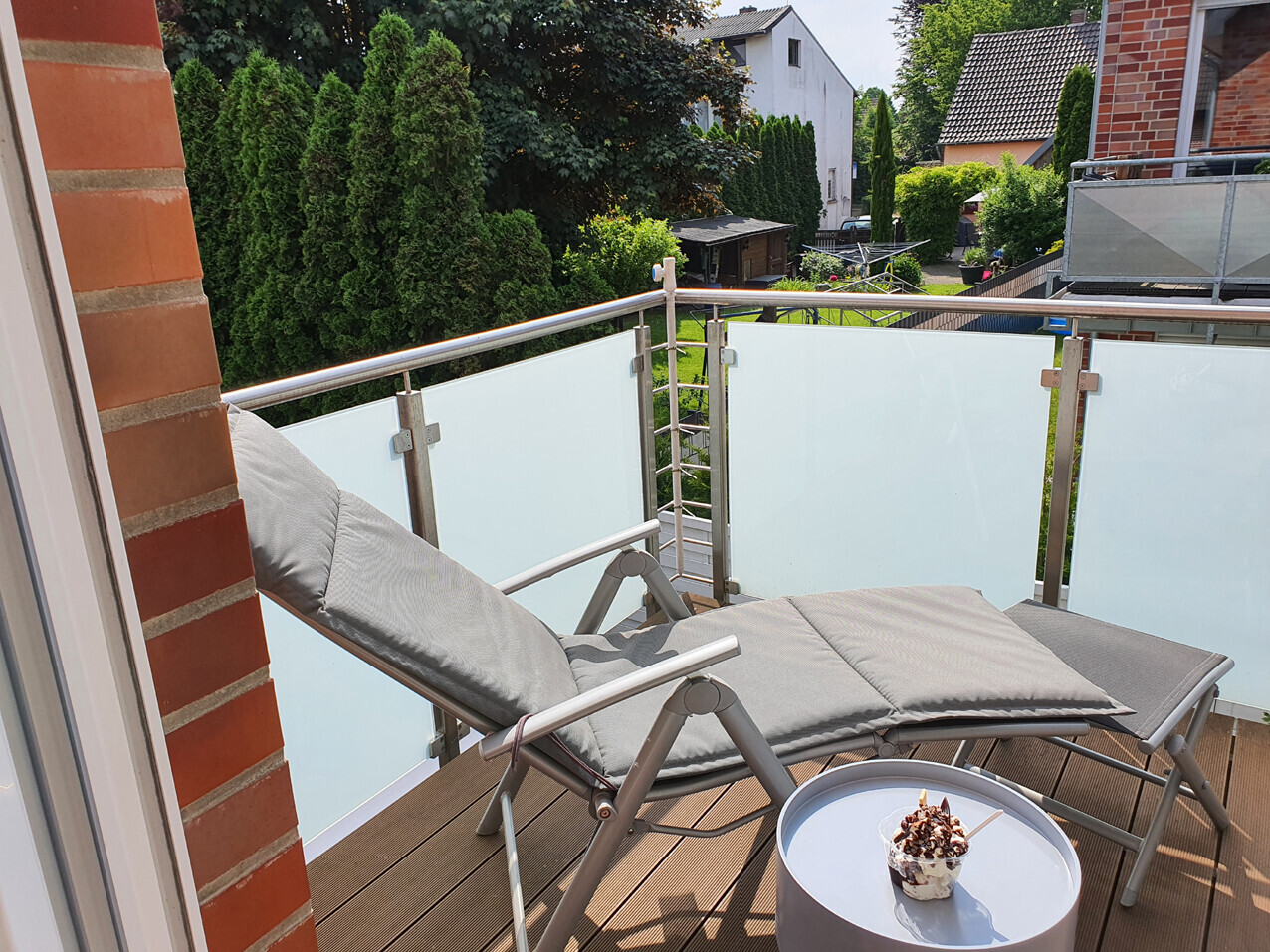 Balkon mit zwei Liegestühlen mit grauen Kissen, Glasgeländer, runder Tisch mit Dessert, Gartenblick.
