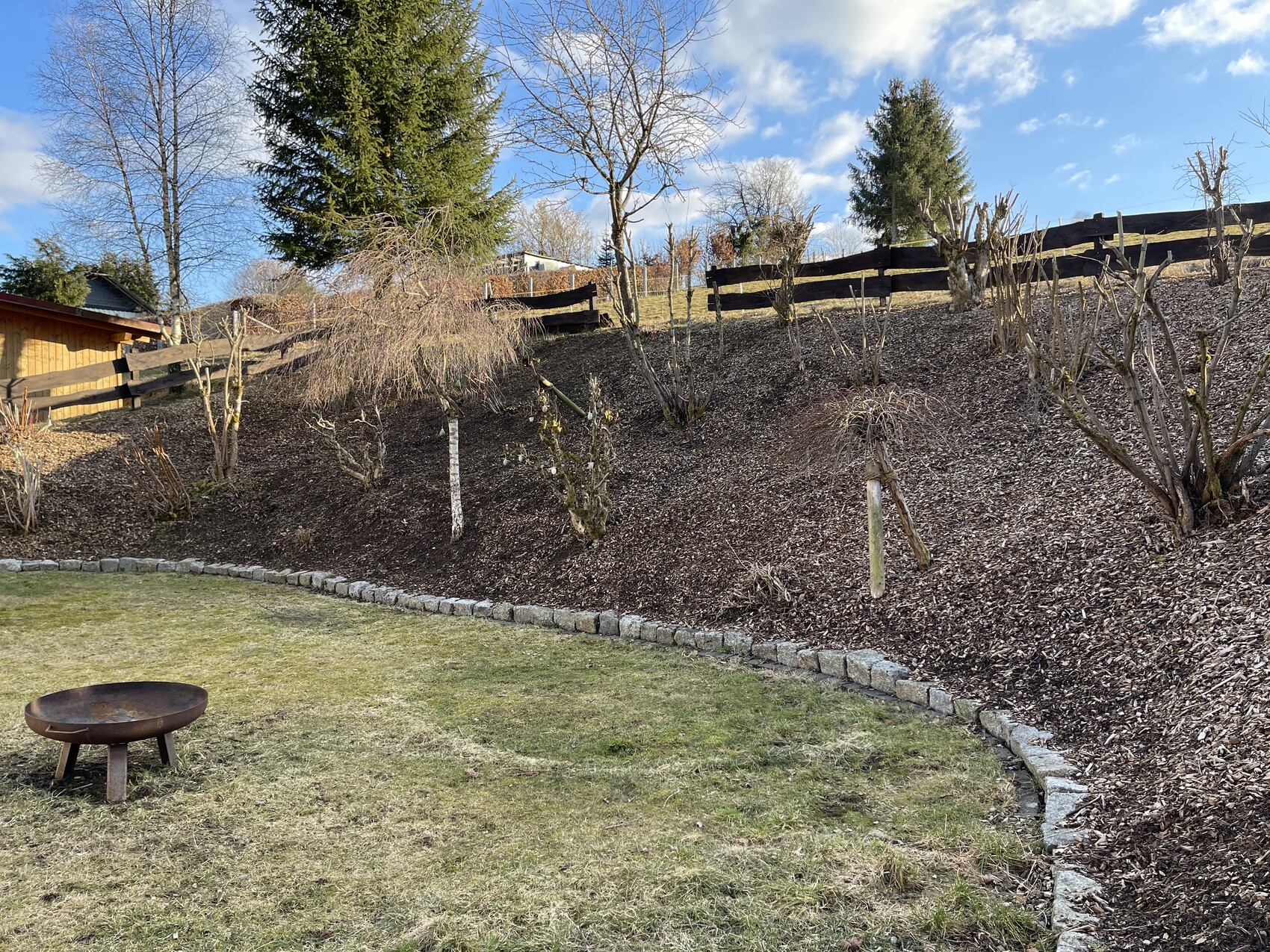 Gartenhang mit Mulch, kahlen Sträuchern, Holzzaun am Hang und Feuerschale im Vordergrund