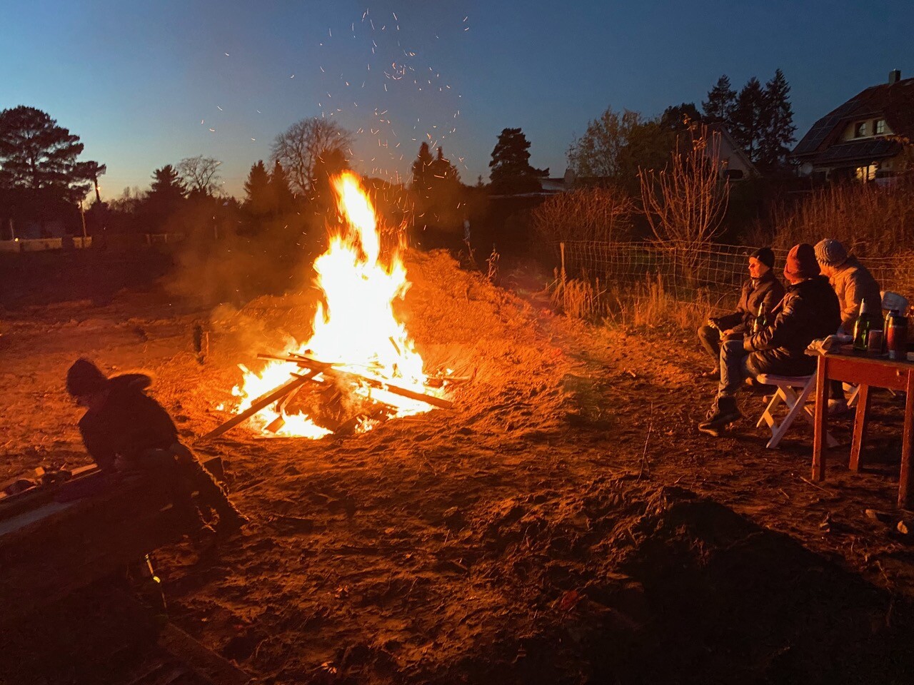 Gruppe sitzt am Lagerfeuer im Freien, Funken sprühen, Abenddämmerung, Tisch mit Getränken rechts.