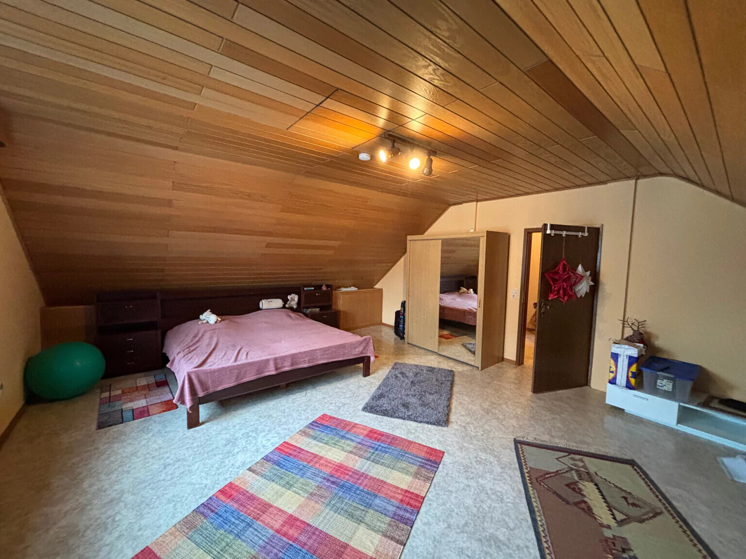 Dachgeschosszimmer mit Holzdecke, Bett mit pinkem Bezug, Kleiderschrank, Spiegel, Teppiche.