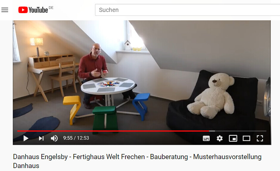 Mann im roten Hemd sitzt am runden Spieltisch; bunte Stühle, Teddy auf schwarzem Sessel.