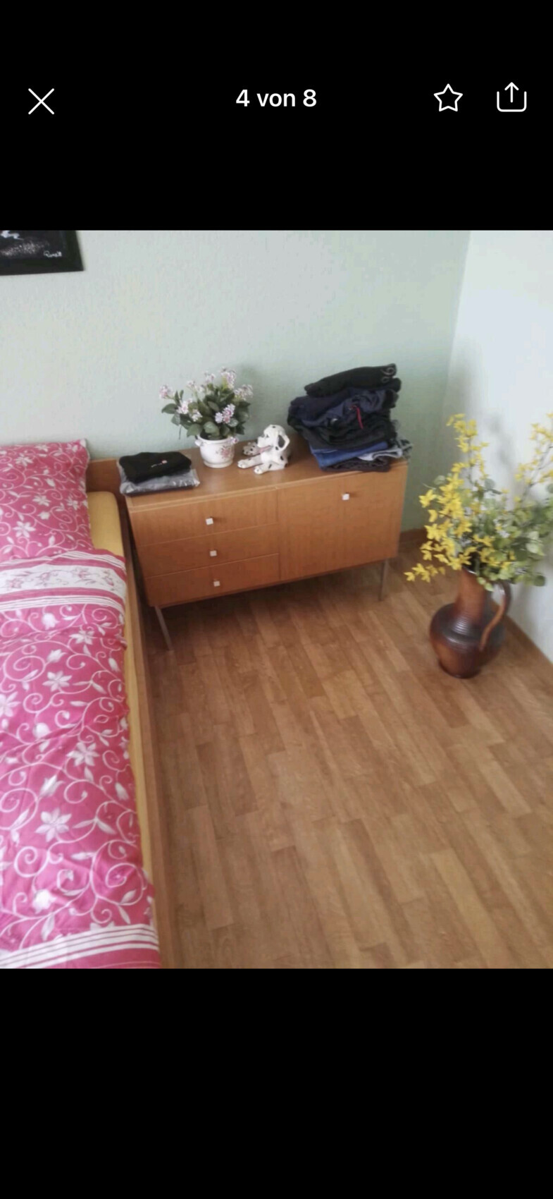 Schlafzimmer-Ecke: Bett links, Holzmöbel mit Kleiderstapeln, Blumen, Hundestatuette Gelbblüten-Vase.