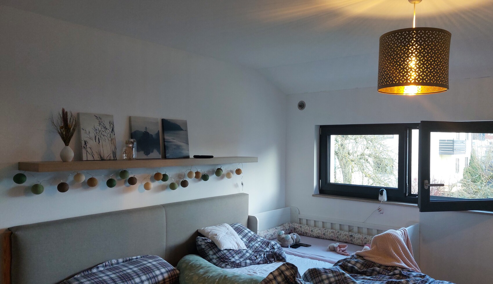 Schlafzimmer mit Bett, Plaids, Wandregal und bunten Kugelkette-Deko; offenes Fenster, warmes Licht.