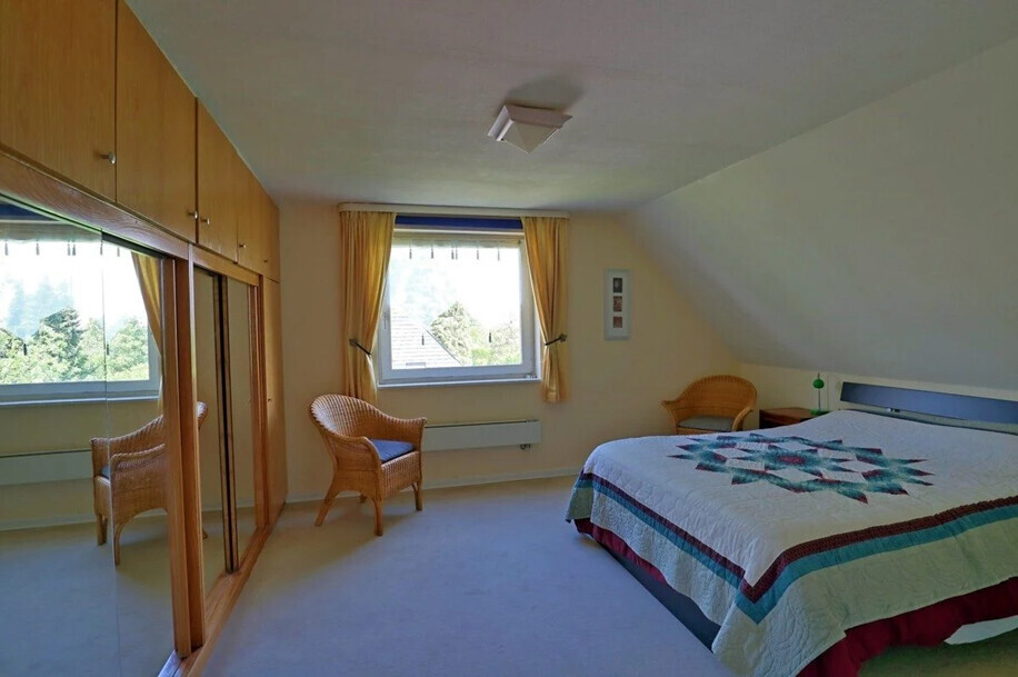 Schlafzimmer mit Quilt-Bett, zwei Korbsessel, Kleiderschrank und Fenster mit Vorhängen.