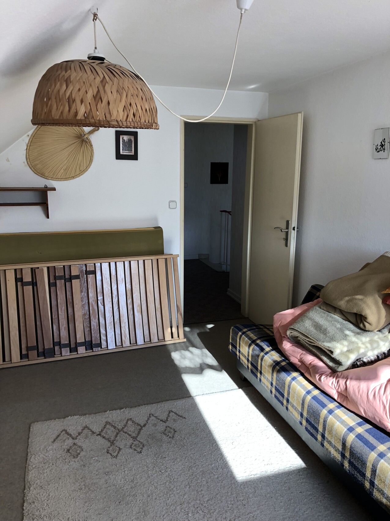 Helles Zimmer mit Bettgestell, gestapelten Decken, Weidenlampe und offener Tür zum Flur.
