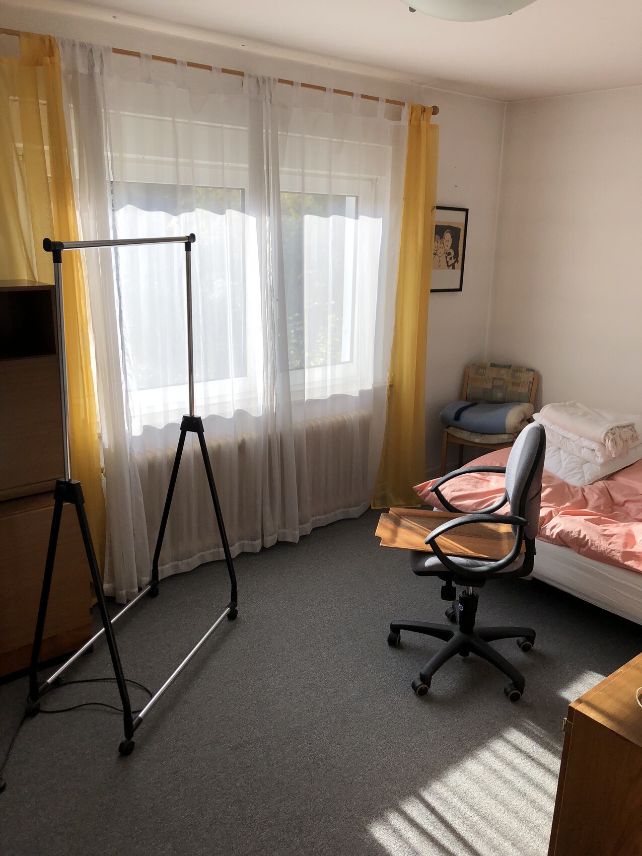 Schlafzimmer mit Fenster, gelben Vorhängen, Kleiderständer links, Bürostuhl neben dem Bett.