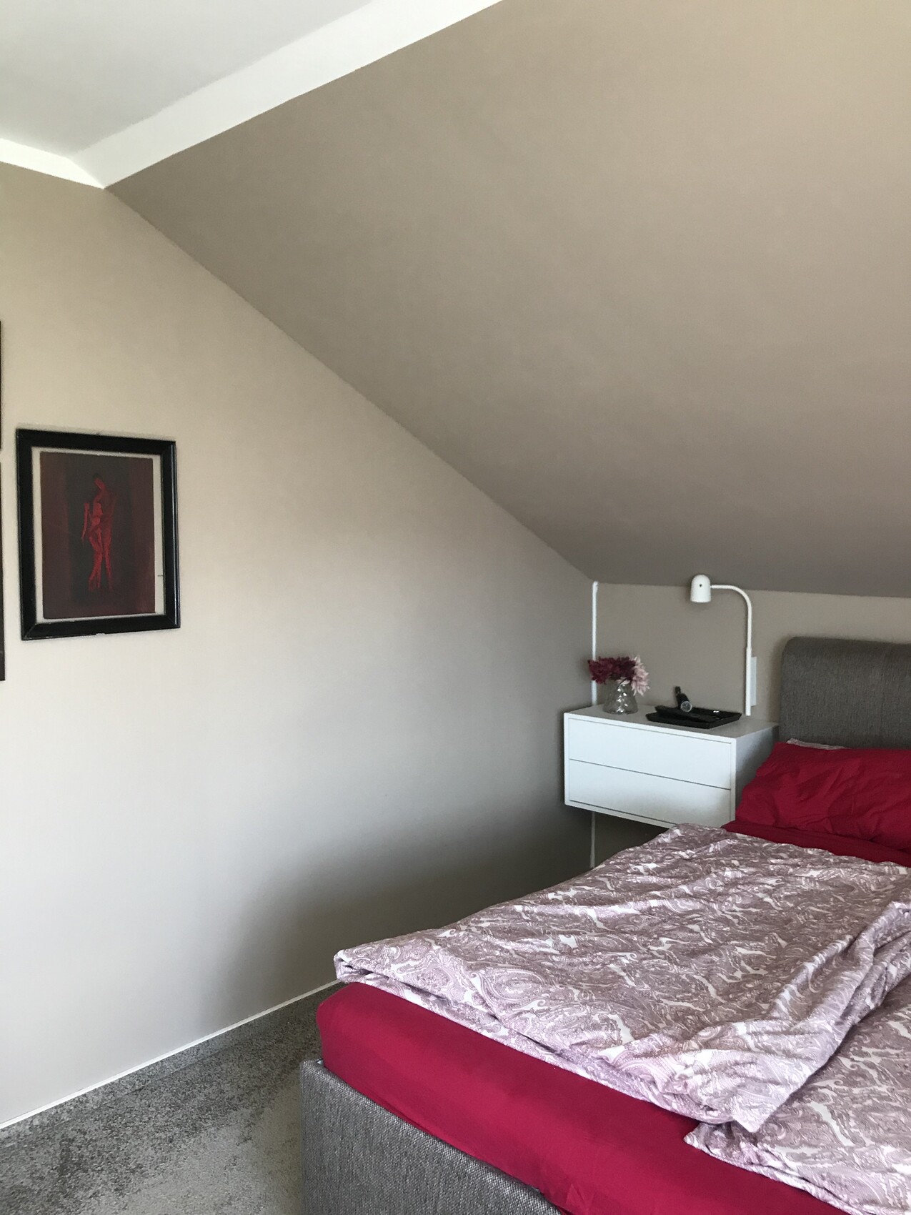 Schlafzimmer mit schräger Decke, rotes Bettzeug, weißer Nachttisch, Lampe, Vase mit Blumen