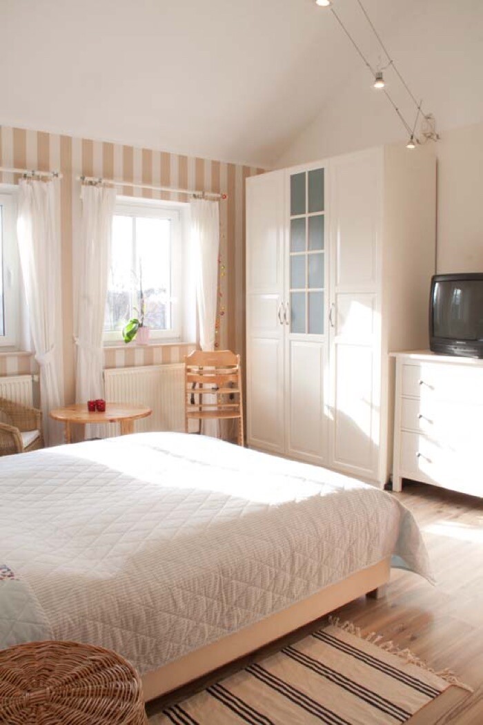 Helles Schlafzimmer mit Bett, Kleiderschrank, TV, Holzfußboden und Fenster mit Vorhängen.